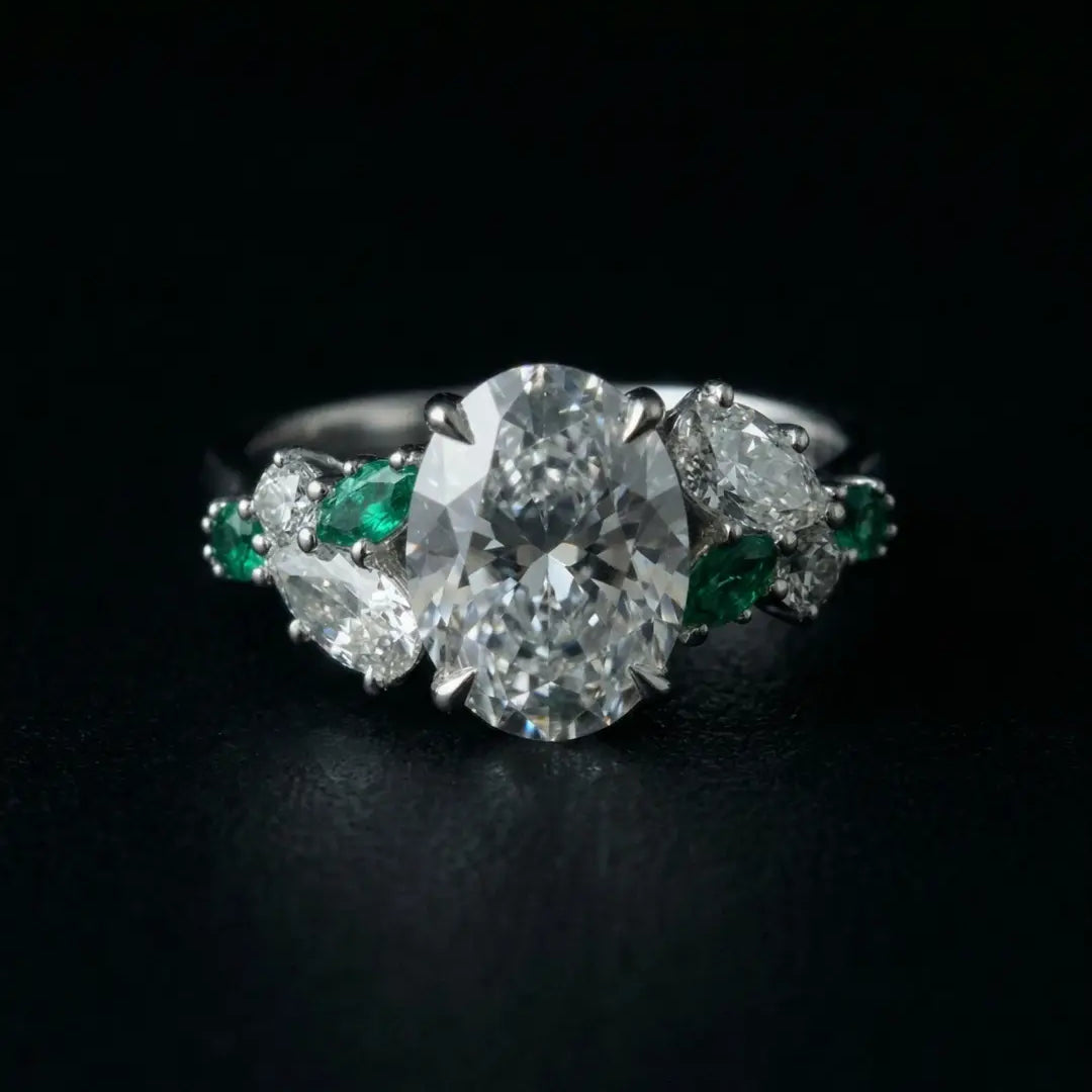 Oval-cut diamond engagement ring in platinum with pear-shaped diamond and emerald side stones, handcrafted by Dimis Designs with heirloom-quality detail.